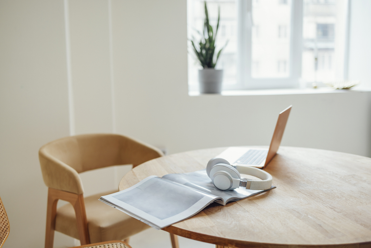 Laptop, headphones and magazine on a round wooden table next to a comfortable beige armchair.
