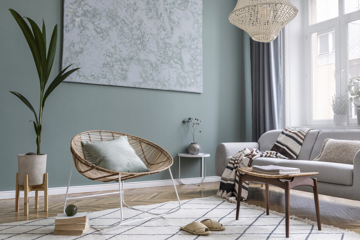 Modern and bohemian composition of interior design with gray sofa, rattan armchair, retro footrest, plaid, pillow, tropical plants, small table and elegant accessories. Stylish home decor. Template.